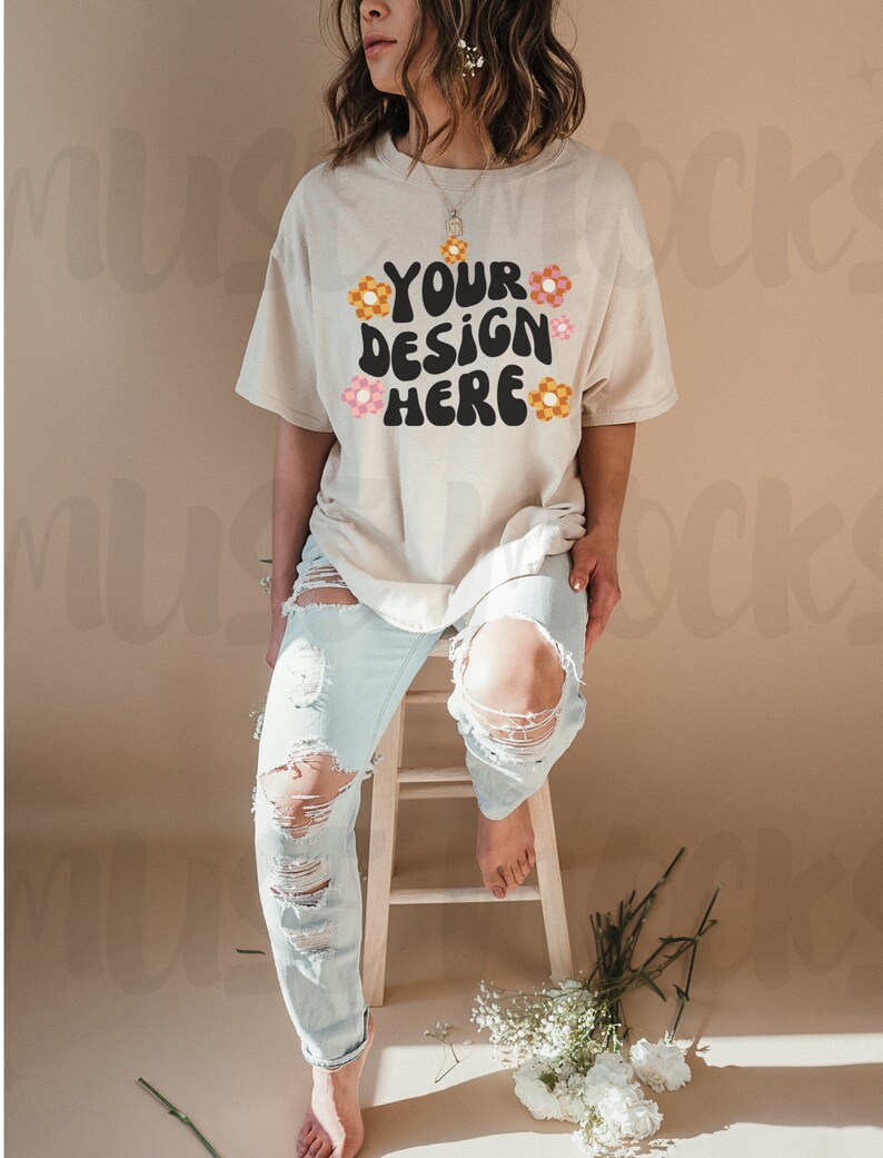 May include: A beige t-shirt with the text "YOUR DESIGN HERE" in black, decorated with orange and pink flowers. The model wears light blue ripped jeans and sits on a wooden stool. White flowers and greenery are on the floor.