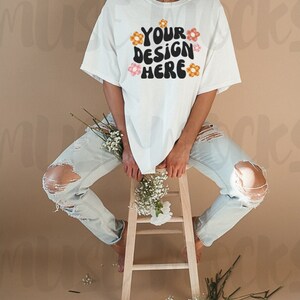 May include: A white t-shirt with the text "YOUR DESIGN HERE" in black, with orange and pink flower accents. The model is wearing light blue ripped jeans and sitting on a wooden stool. White flowers are placed around the model.