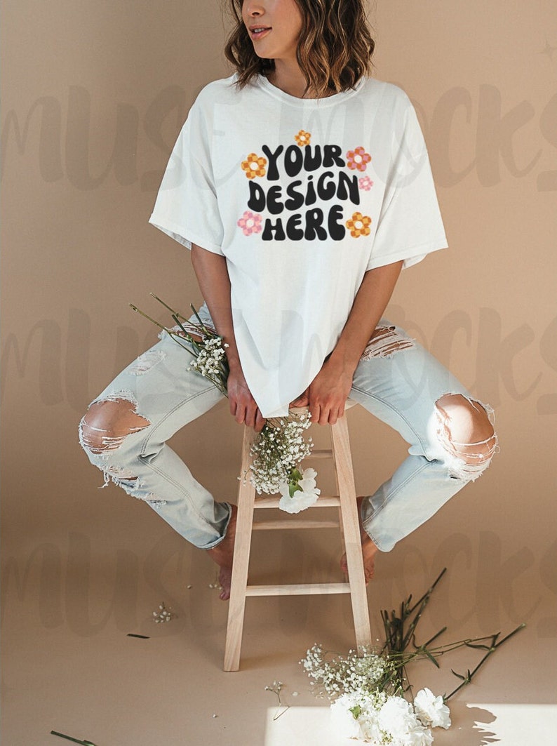 May include: A white t-shirt with the text "YOUR DESIGN HERE" in black, with orange and pink flower accents. The model is wearing light blue ripped jeans and sitting on a wooden stool. White flowers are placed around the model.