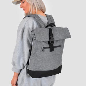 Può includere: Zaino grigio con base nera e chiusura arrotolabile assicurata da una fibbia nera. Lo zaino ha una tasca frontale con cerniera e spallacci regolabili. Viene indossato un berretto da baseball beige.