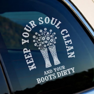 May include: A white vinyl decal applied to a car window featuring a graphic of two cowboy boots filled with a bouquet of daisies. The text surrounding the illustration reads, "KEEP YOUR SOUL CLEAN AND YOUR BOOTS DIRTY" in a bold, arched font.