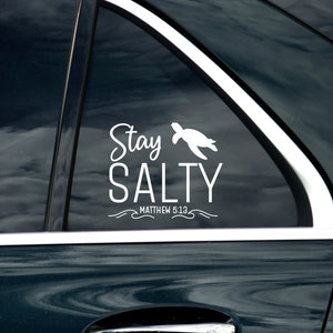 Puede incluir: Calcomanía de vinilo blanco aplicada a la ventana de un coche. La calcomanía presenta las palabras "Stay Salty" en una fuente cursiva, con un gráfico de tortuga y el texto "Matthew 5:13" debajo. El coche es azul oscuro.