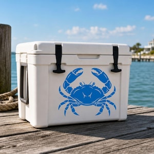 May include: A white rectangular cooler featuring a blue graphic of a crab on the front panel. The cooler has two black rubber latches on the lid and a black handle on the side, designed for keeping items cold while outdoors or near the water.