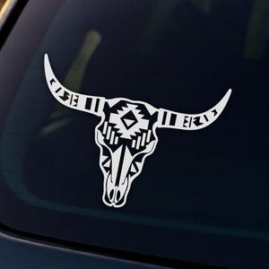 Puede incluir: Calcomanía de vinilo blanco de una calavera de toro con patrones geométricos, aplicada a la ventana trasera de un vehículo negro. El fondo muestra un lago y árboles al atardecer.