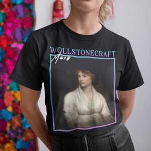 Pode incluir: Camiseta preta com um retrato de Mary Wollstonecraft com um vestido branco. O texto "WOLLSTONECRAFT Mary" é impresso em uma fonte retrô acima do retrato.