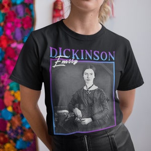 Pode incluir: Camiseta preta com uma estampa gráfica de Emily Dickinson, uma famosa poetisa. O gráfico está em um estilo retrô com uma borda roxa e azul. O texto "Dickinson" está em negrito, em branco e "Emily" está em itálico, em branco.