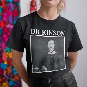 Pode incluir: Uma camiseta preta com um gráfico branco de um retrato de Emily Dickinson e o texto "Dickinson" acima do retrato.