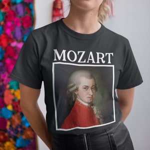 May include: A black t-shirt with a white print of a portrait of Mozart. The portrait is in a white frame and the word "MOZART" is printed above the frame.