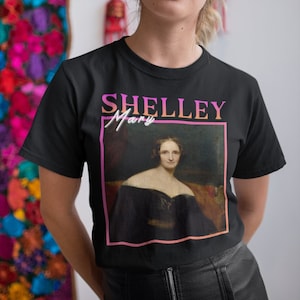 Pode incluir: Camiseta preta com um gráfico rosa e laranja do retrato de Mary Shelley. O texto "SHELLEY Mary" está impresso acima do retrato.