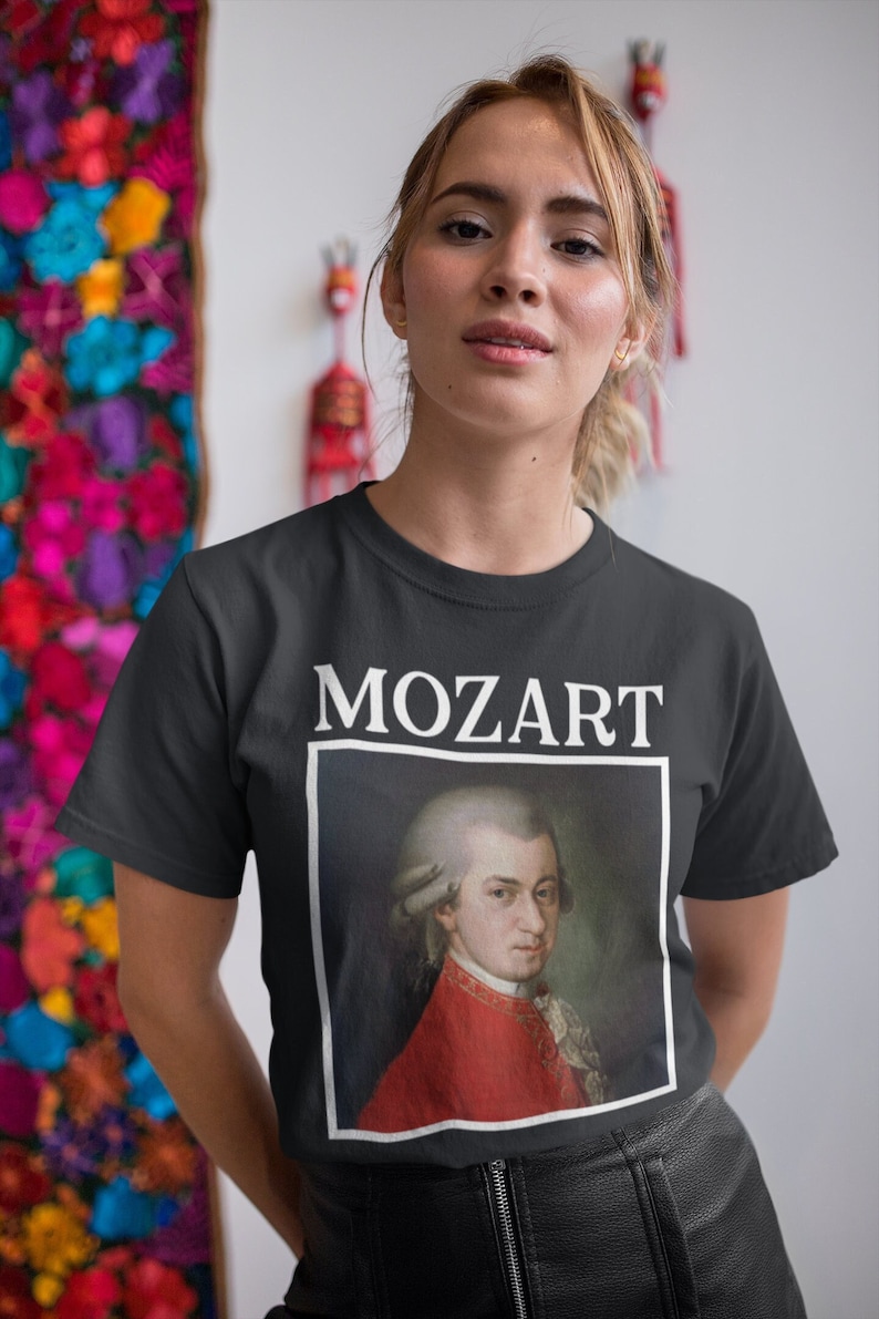 May include: A black t-shirt with a white print of a portrait of Mozart. The portrait is in a white frame and the word "MOZART" is printed above the frame.