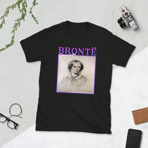 Pode incluir: Camiseta preta com um retrato de Charlotte Bront&euml; em uma moldura branca. O texto "BRONT&Euml;" est&aacute; acima do retrato em letras roxas e rosas. O texto "Charlotte" est&aacute; abaixo do retrato em letras brancas.