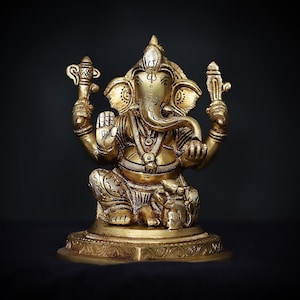 Op de afbeelding: Een gouden metalen beeld van de hindoe&iuml;stische god Ganesha. Het beeld toont Ganesha zittend met zijn slurf omhoog en een klein voorwerp in zijn hand. Het beeld is versierd met ingewikkelde details en staat op een voetstuk.
