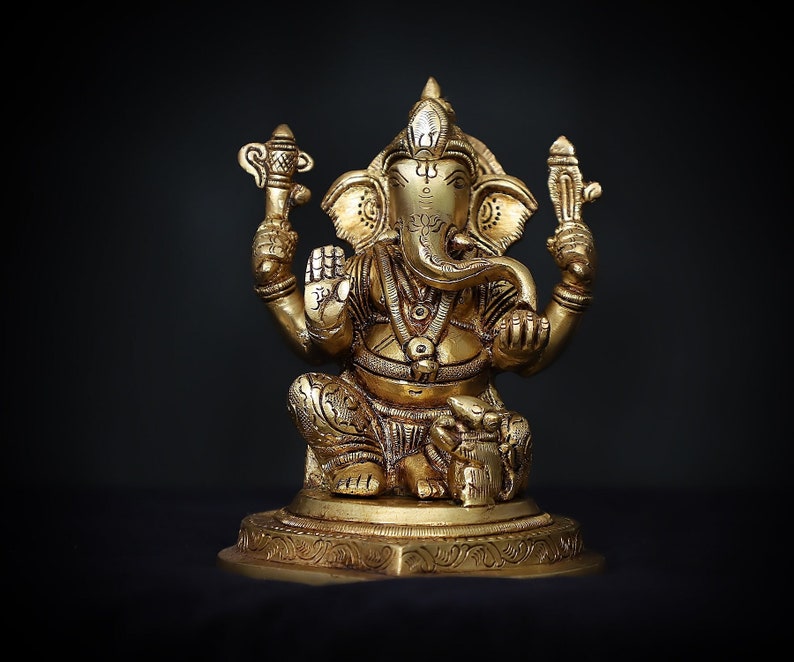 Op de afbeelding: Een gouden metalen beeld van de hindoe&iuml;stische god Ganesha. Het beeld toont Ganesha zittend met zijn slurf omhoog en een klein voorwerp in zijn hand. Het beeld is versierd met ingewikkelde details en staat op een voetstuk.