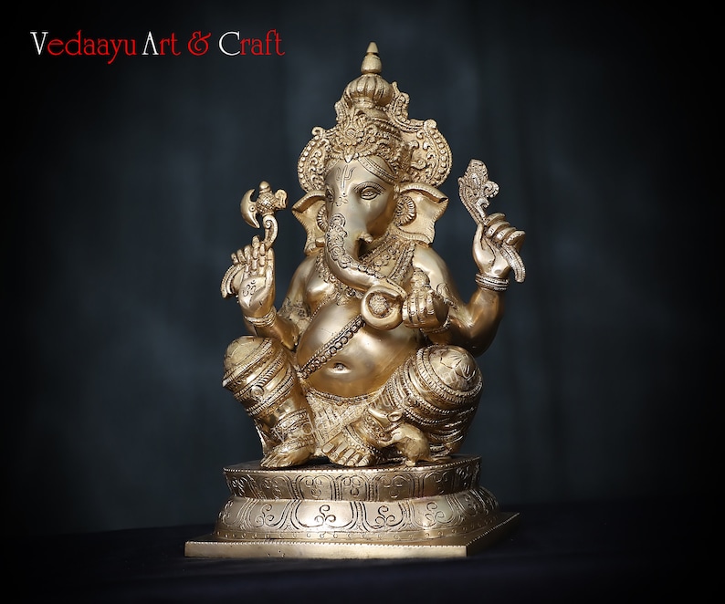 May include: A gold-colored brass statue of Ganesha, the Hindu god of wisdom and prosperity, sitting on a pedestal with intricate carvings. The statue is detailed with ornate jewelry and a trunk.
