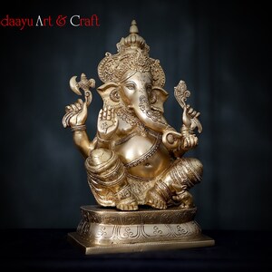 May include: A gold-colored brass statue of Ganesha, the Hindu god of wisdom and prosperity. The statue is sitting on a base with intricate carvings. The statue is holding a small axe in one hand and a sweet in the other.