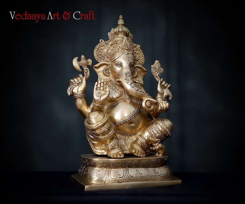 May include: A gold-colored brass statue of Ganesha, the Hindu god of wisdom and prosperity. The statue is sitting on a base with intricate carvings. The statue is holding a small axe in one hand and a sweet in the other.