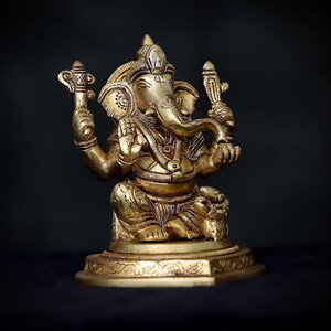 Op de afbeelding: Een gouden metalen beeld van de hindoe&iuml;stische god Ganesha. Het beeld toont Ganesha zittend met zijn slurf omhoog en een klein voorwerp in zijn hand. Het beeld is versierd met ingewikkelde details en staat op een voetstuk.