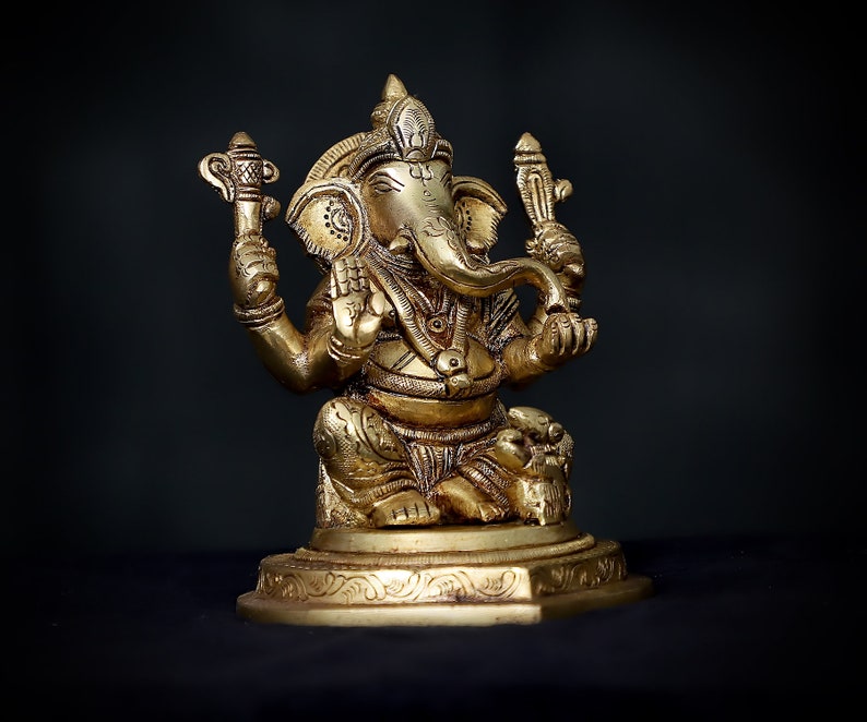 Op de afbeelding: Een gouden metalen beeld van de hindoe&iuml;stische god Ganesha. Het beeld toont Ganesha zittend met zijn slurf omhoog en een klein voorwerp in zijn hand. Het beeld is versierd met ingewikkelde details en staat op een voetstuk.