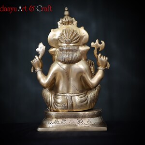 May include: A gold-colored brass statue of Ganesha, the Hindu god of wisdom and prosperity, sitting with his trunk raised and holding a sweet in his hand. The statue is on a base with intricate carvings.