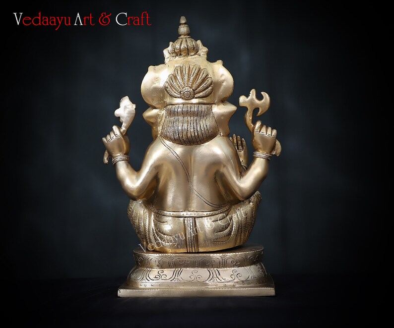 May include: A gold-colored brass statue of Ganesha, the Hindu god of wisdom and prosperity, sitting with his trunk raised and holding a sweet in his hand. The statue is on a base with intricate carvings.
