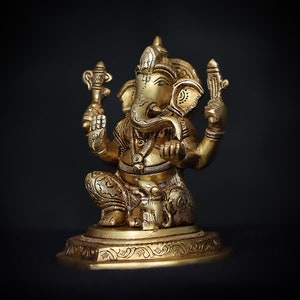 Op de afbeelding: Een gouden beeld van Ganesha, de hindoe&iuml;stische god van wijsheid en voorspoed, zittend op een basis met ingewikkelde houtsnijwerk. Het beeld is gedetailleerd met sierlijke ontwerpen en heeft een slurf, grote oren en &eacute;&eacute;n slagtand.