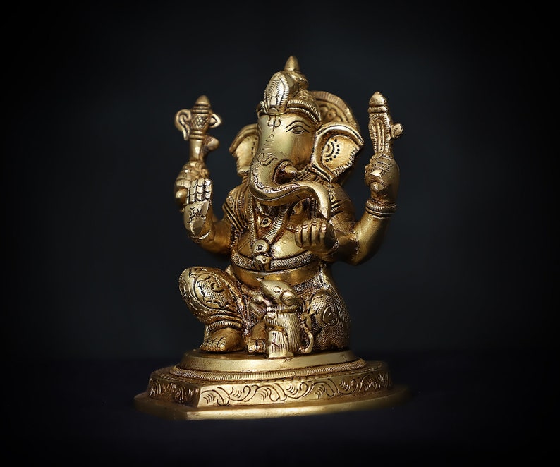 Op de afbeelding: Een gouden beeld van Ganesha, de hindoe&iuml;stische god van wijsheid en voorspoed, zittend op een basis met ingewikkelde houtsnijwerk. Het beeld is gedetailleerd met sierlijke ontwerpen en heeft een slurf, grote oren en &eacute;&eacute;n slagtand.