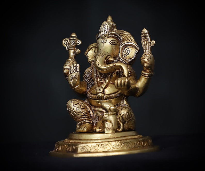 Op de afbeelding: Een gouden beeld van Ganesha, de hindoe&iuml;stische god van wijsheid en voorspoed, zittend op een basis met ingewikkelde details. Het beeld is gemaakt van metaal en heeft een gedetailleerd ontwerp.