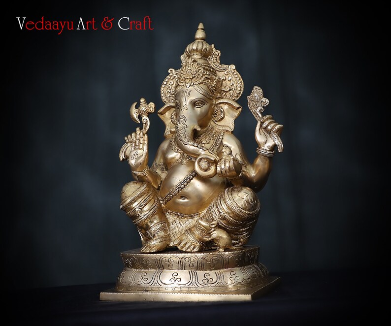 May include: A gold-colored brass statue of Ganesha, the Hindu god of wisdom and prosperity, sitting on a pedestal with intricate carvings. The statue is detailed with ornate jewelry and a trunk.