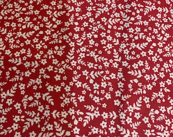May include: Red fabric with a repeating pattern of small white flowers and leaves. The floral design covers the entire surface, creating a dense and visually appealing texture. The fabric is likely suitable for various sewing and crafting projects.
