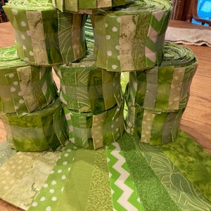 May include: A stack of green fabric rolls with different patterns, including polka dots, stripes, and swirls. The rolls are arranged in a pyramid shape and are surrounded by flat pieces of fabric in various green shades.