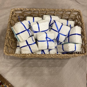 May include: A wicker basket filled with white fabric squares tied with blue ribbon. The squares are stacked and arranged in a random pattern.