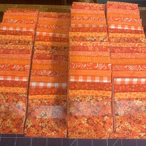 May include: Stacks of orange fabric squares in various patterns, including floral, gingham, and geometric designs. The fabric pieces are arranged in neat rows, showcasing a variety of shades from light to dark orange. Ideal for quilting or crafting projects.