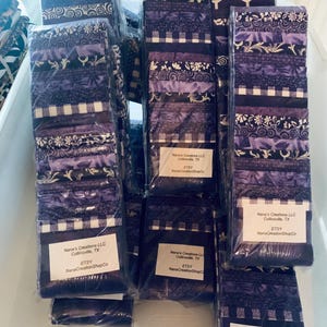 May include: Stacks of fabric bundles in shades of purple, featuring various patterns like floral, gingham, and abstract designs. Each bundle is wrapped in clear plastic. The labels read "Nana's Creations LLC, Collinsville, TX, ETSY NanaCreationShopCo."