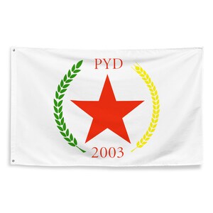 May include: White flag featuring a central red star encircled by green and yellow wheat stalks. The flag has the letters "PYD" in red above the star and the number "2003" below it. The flag is rectangular with grommets along the top edge.