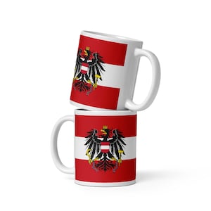 May include: Two white ceramic mugs stacked vertically, each featuring a horizontal red and white striped flag pattern. Centered on each mug is a black eagle emblem with a gold crown, holding a gold hammer and sickle, with a red and white shield.
