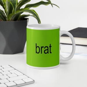Può includere: Una tazza in ceramica bianca con uno sfondo verde brillante e la parola "brat" stampata in nero.