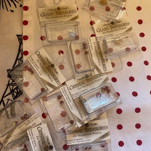 May include: A pile of clear plastic packages containing small red and gold colored glass beads. The packages are labeled "Crystal Treasures" and "Glass Treasures" for needlework projects. The packages are made in Austria and Germany.