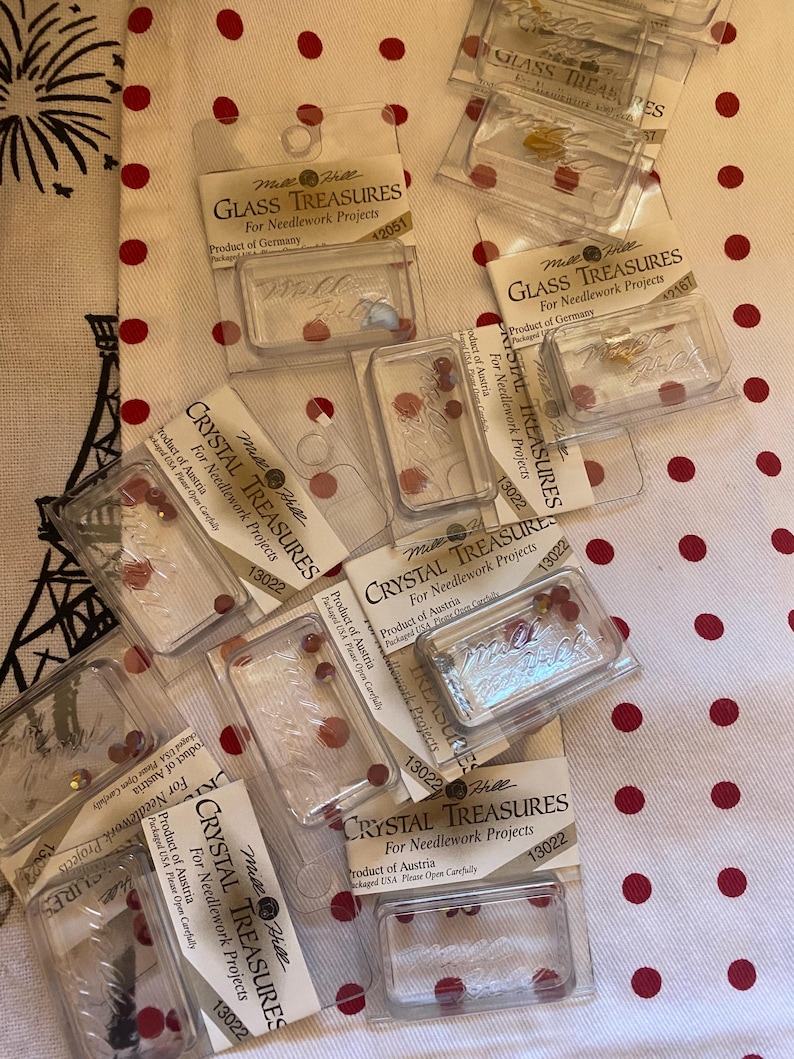 May include: A pile of clear plastic packages containing small red and gold colored glass beads. The packages are labeled "Crystal Treasures" and "Glass Treasures" for needlework projects. The packages are made in Austria and Germany.