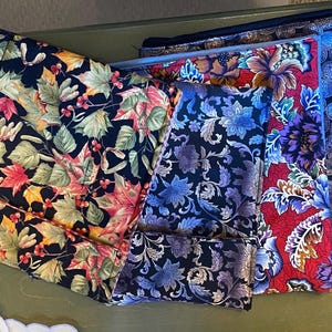 Può includere: Una collezione di pochette in tessuto con vari disegni floreali e fantasia. Le pochette presentano uno sfondo scuro con foglie colorate, fiori e motivi a moneta. I colori includono rosso, blu, viola e oro.