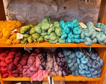 Könnte beinhalten: Ausstellung farbenfroher Garnknäuel auf Holzregalen. Das Garn ist in einem Spektrum von Farben erhältlich, darunter Gelb, Grün, Blau, Rot, Rosa und Lila. Jedes Knäuel ist ordentlich gebündelt und zeigt die Textur und Lebendigkeit des Materials.