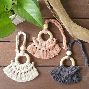 May include: Three macrame car air fresheners in cream, dusty rose, and charcoal grey. Each features a wooden ring and bead, with a knotted design and fringe. The air fresheners hang from a braided cord.