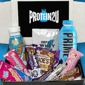 Könnte beinhalten: Eine Box gefüllt mit Protein-Snacks und -Getränken. Die Box enthält ein blaues Himbeer-PRIME Hydration-Getränk, einen Proteinshake, einen Proteinriegel und eine Tüte Proteinpulver. Die Box enthält außerdem eine Auswahl an Proteinriegeln, darunter einen Schokoladen-Chip-Cookie-Teig-Riegel, einen Erdnussbutter-Riegel und einen Schokoladenriegel.