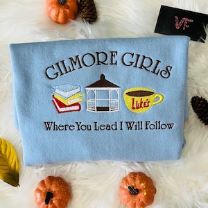 May include: Light blue sweatshirt with the text "Gilmore Girls" embroidered in brown. The sweatshirt also features embroidered images of a stack of books, a gazebo, and a coffee cup with the text "Luke's". The text "Where You Lead I Will Follow" is also embroidered on the sweatshirt.