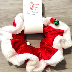 May include: A festive red and white scrunchie with a Santa Claus-inspired design. The scrunchie features a plush white trim and is adorned with small red and green jingle bells. A white tag reads "Made It For U Handmade Scrunchies #208."