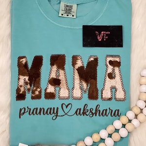 May include: A turquoise blue t-shirt with a brown and white cow print design that says "MAMA" with the text "pranay akshara" underneath.