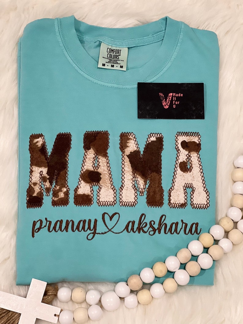 May include: A turquoise blue t-shirt with a brown and white cow print design that says "MAMA" with the text "pranay akshara" underneath.