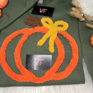 May include: Olive green sweatshirt with a large orange pumpkin design, a brown stem, and a yellow bow. The design is made of a textured, fuzzy material. Two small black rectangles with text are visible. Decorative pumpkins are placed around the sweatshirt.