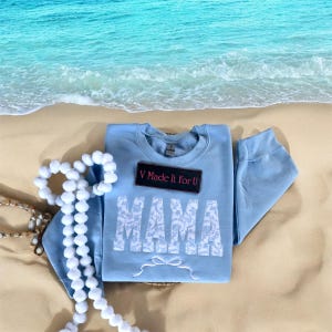 May include: Light blue sweatshirt with a white lace design that spells out "MAMA" with a white bow underneath. The sweatshirt has a black tag that says "V Made It For U".