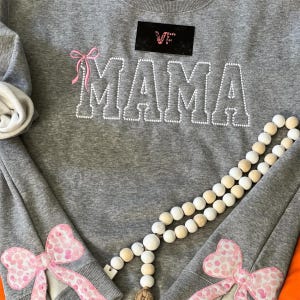 May include: Gray sweatshirt with the word "MAMA" in white rhinestones and pink bows on the sleeves. The sweatshirt has a small black tag with the text "Made It For U".