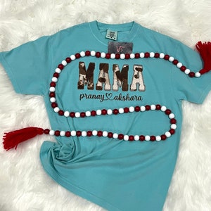 May include: A light blue t-shirt with a brown and white "MAMA" graphic with a cow print design. The shirt also has the text "pranay akshara" in brown lettering with a heart symbol between the words.